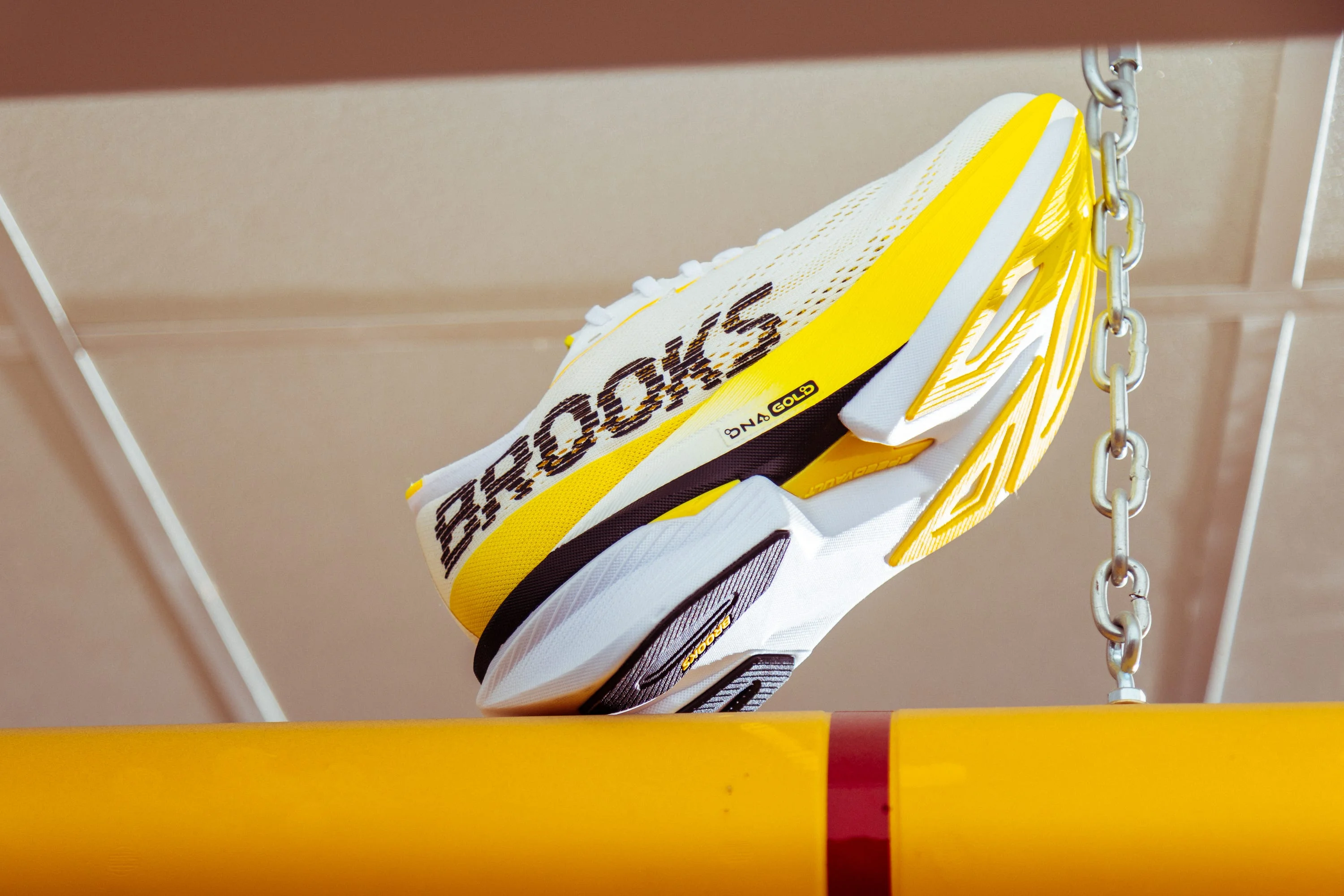 Yellow and white running shoe with "Brooks" branding, displayed hanging above a yellow bar.