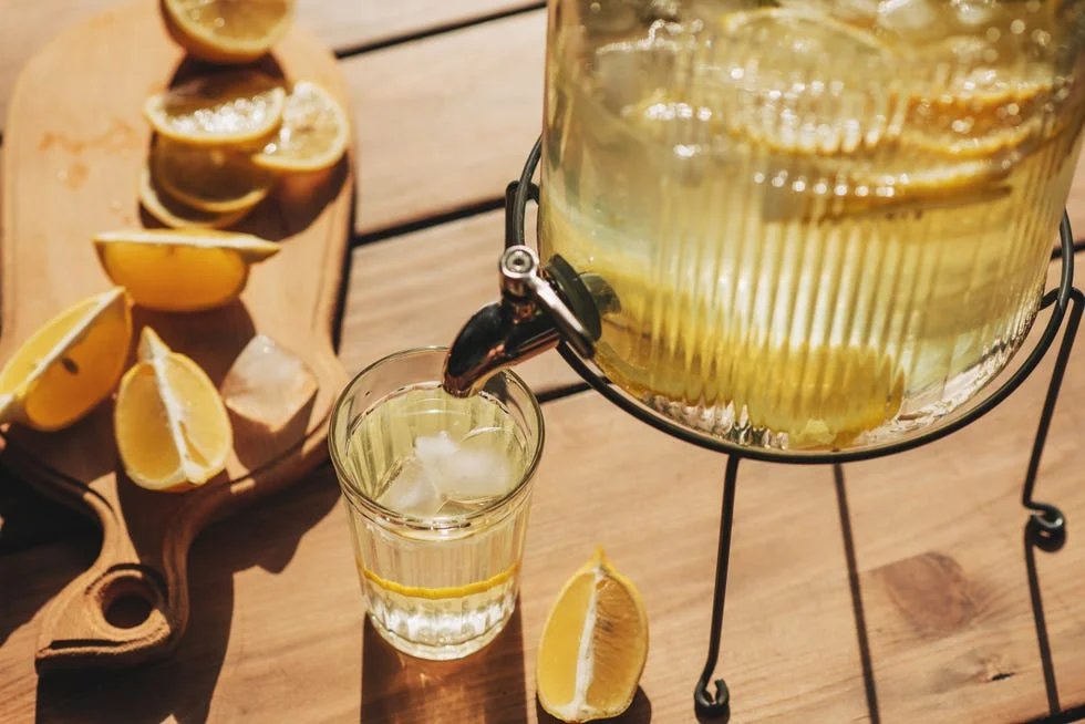 Lemonade with ice in a pitcher and glass on a wooden table with fruit and crushed ice outside with a lemon field in the background on a sunny day. Front view. Horizontal composition.