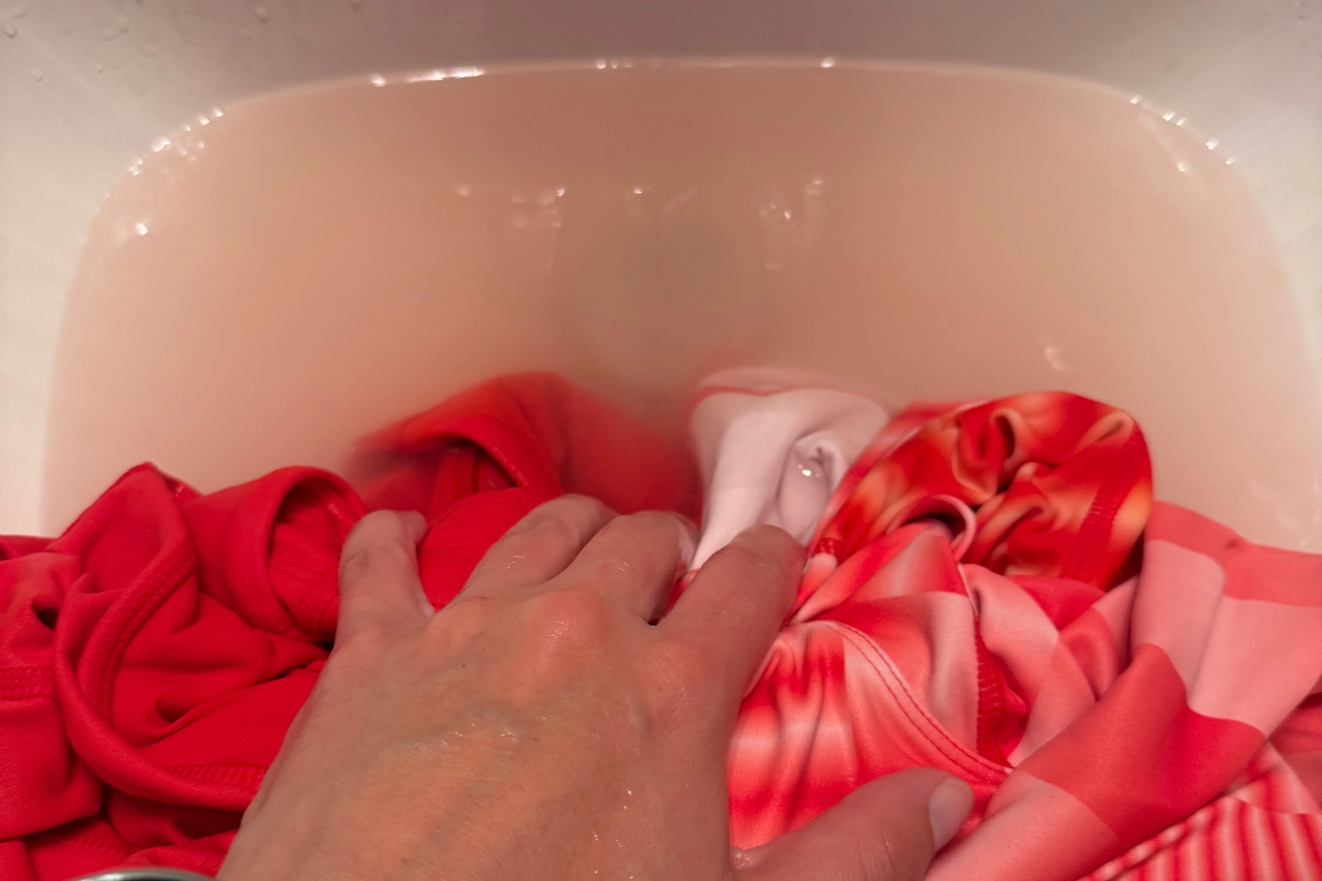 A hand submerged in a sink filled with water and red and pink clothing items.