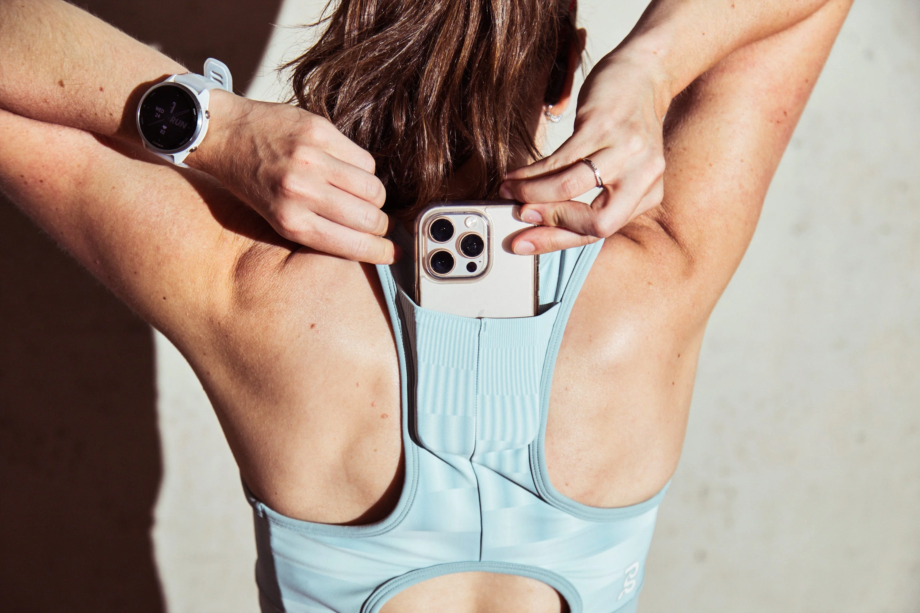 A person securing a smartphone in the back pocket of a light blue athletic top while wearing a fitness watch.