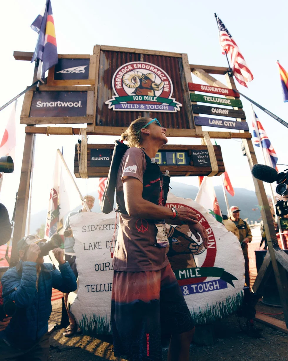 Runner celebrating at the Hardrock Endurance Run finish line, surrounded by flags and photographers.
