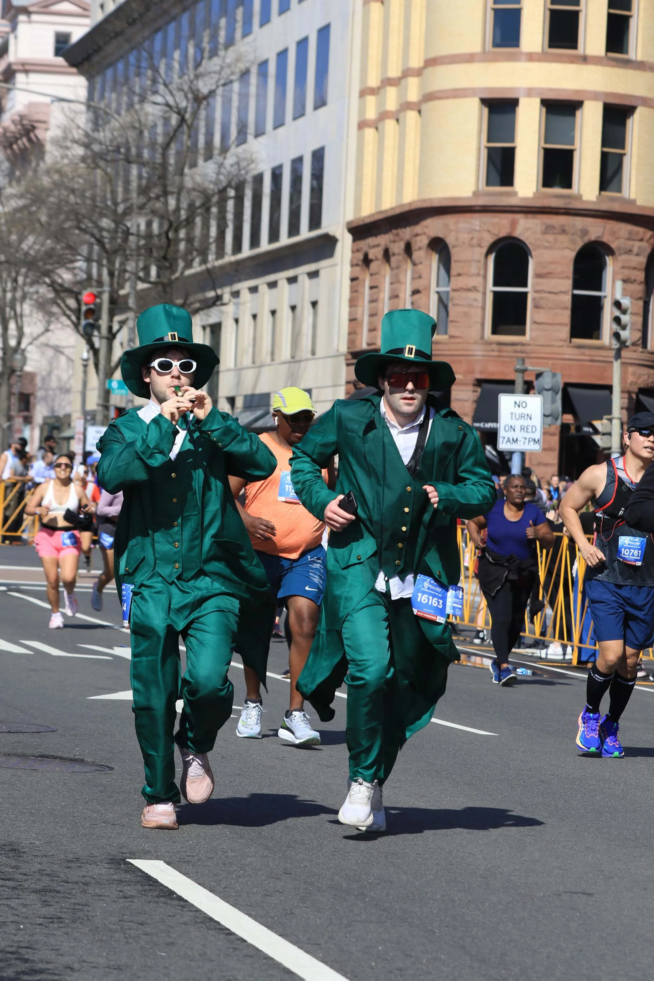 Two runners dressed as leprechauns participate in a lively street race alongside other competitors.
