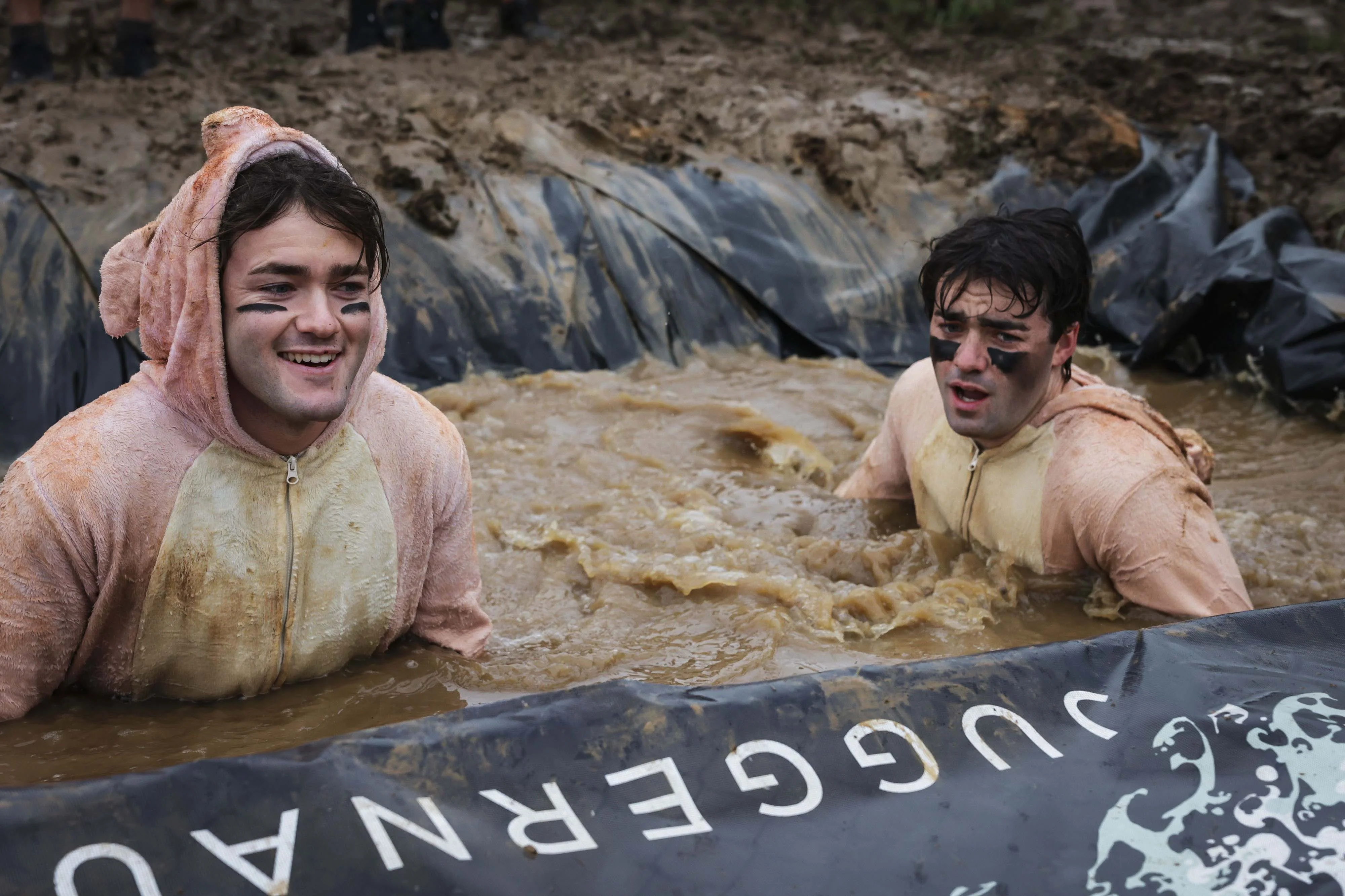 Two men in animal costumes playfully splashing in muddy water at an outdoor event.