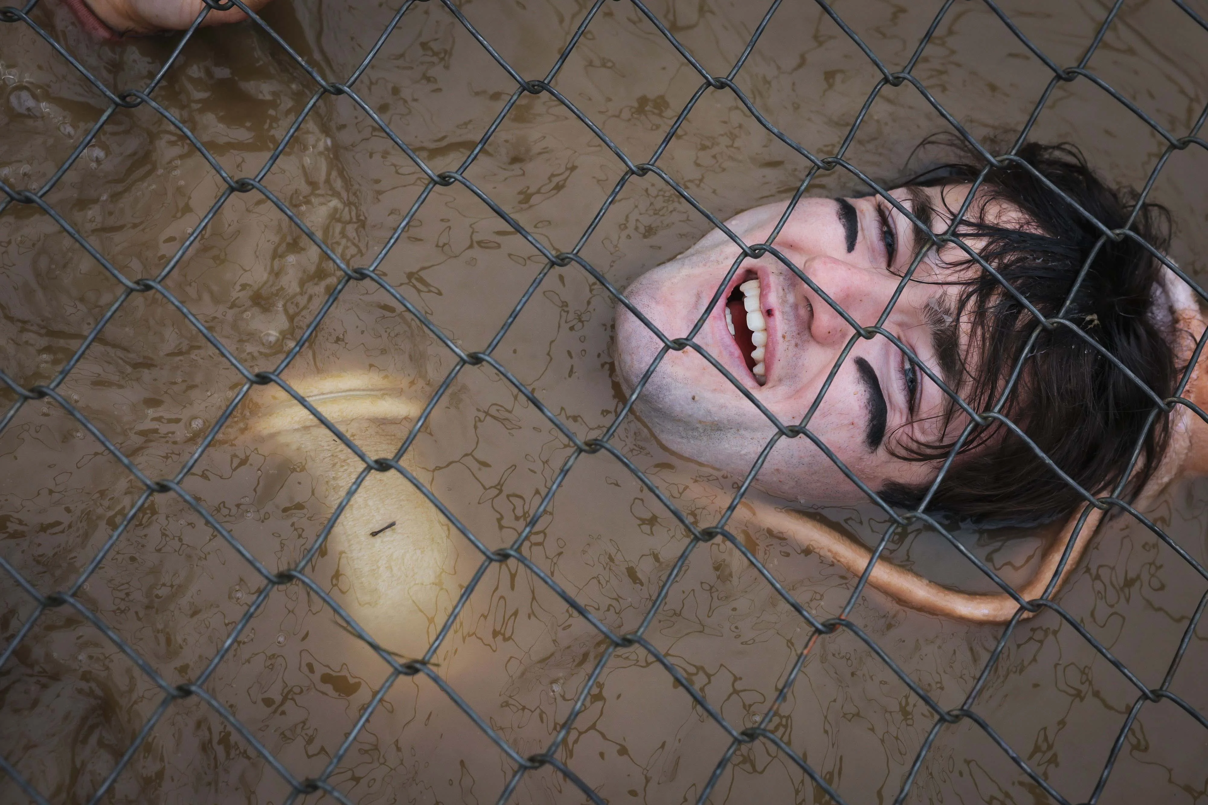 A smiling person submerged in muddy water, partially covered by a mesh fence.