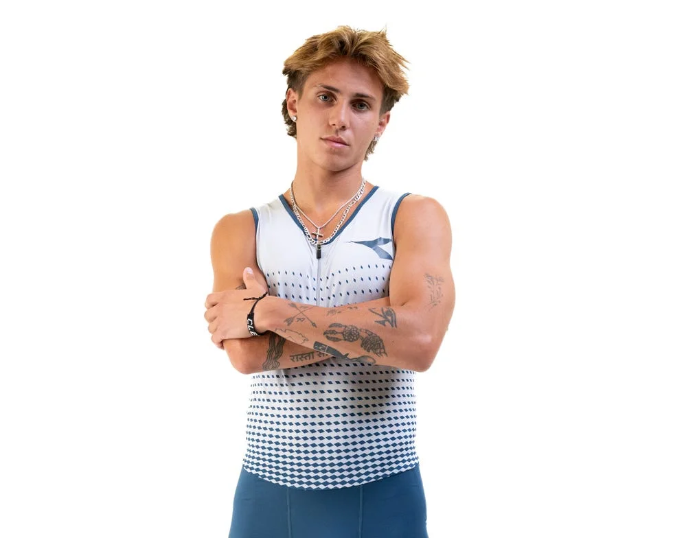 Athlete in a sleeveless athletic top with arms crossed, showcasing tattoos and accessories against a plain background.