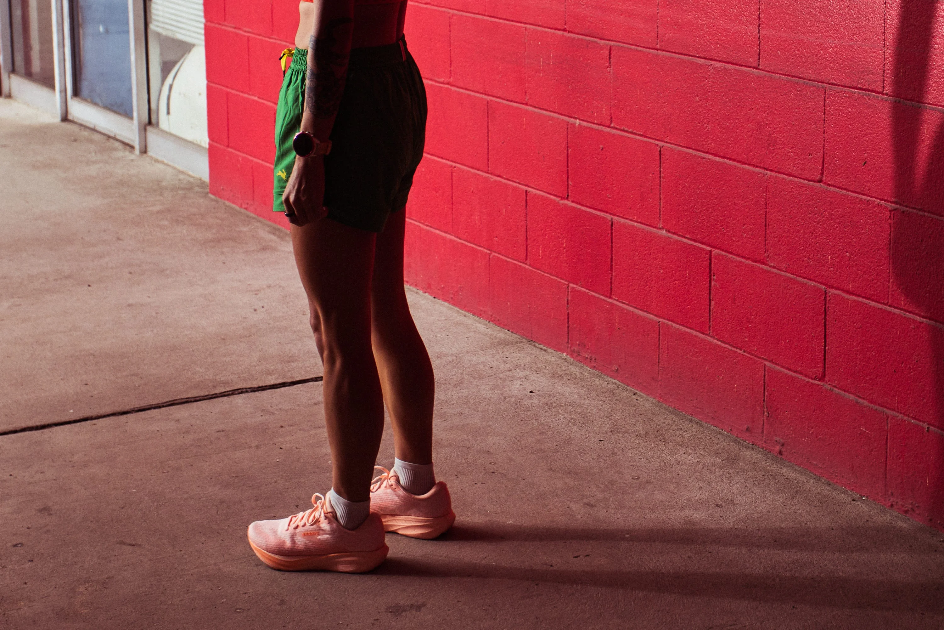 A person standing in sporty attire with shorts and sneakers against a red wall.
