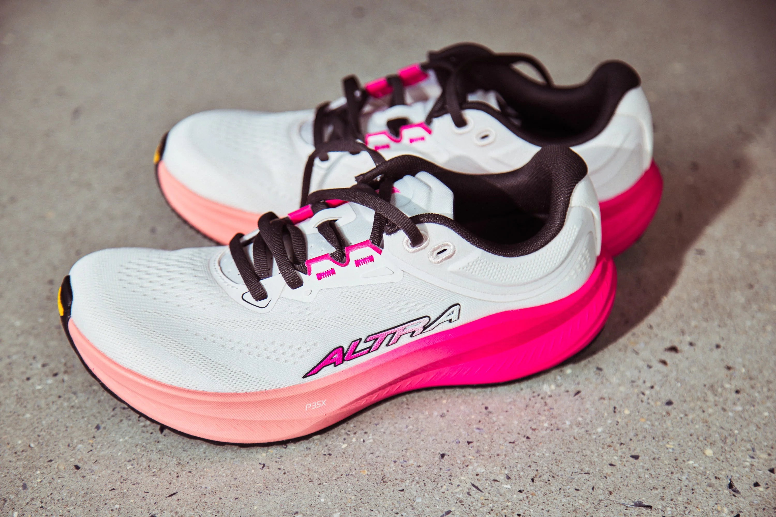 White and pink athletic shoes with black laces and Altra branding on a gray surface.