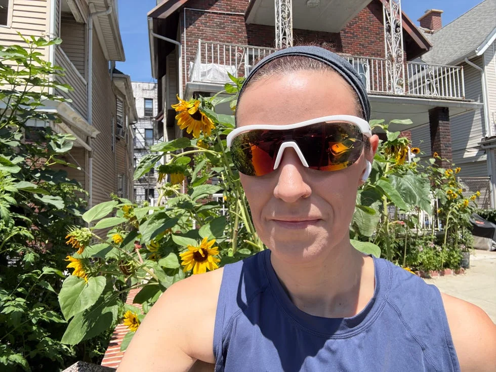 Person wearing sunglasses and a sleeveless top, standing among sunflowers in a sunny outdoor setting.