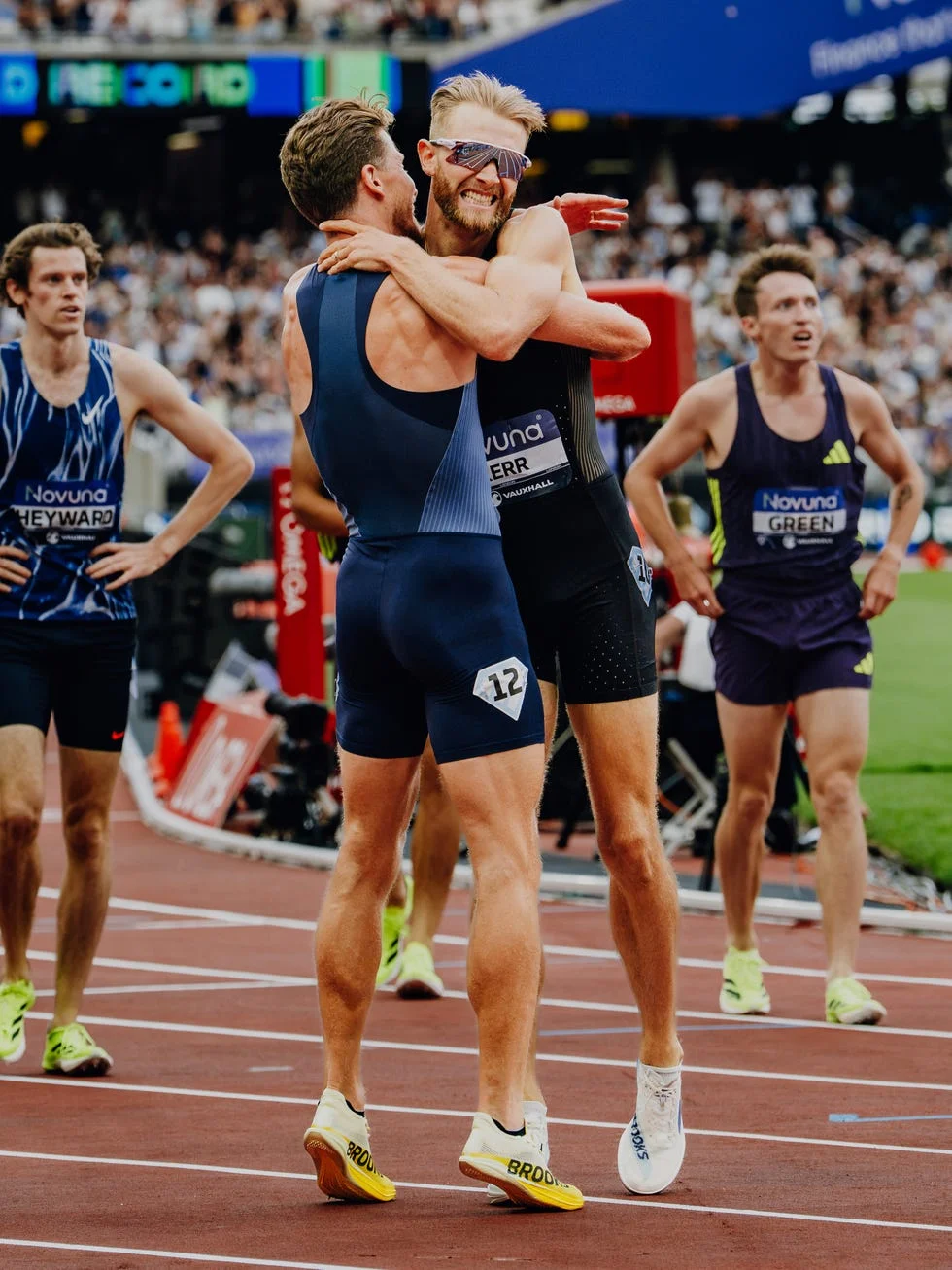Two athletes embrace joyfully after a race, with other runners in the background.