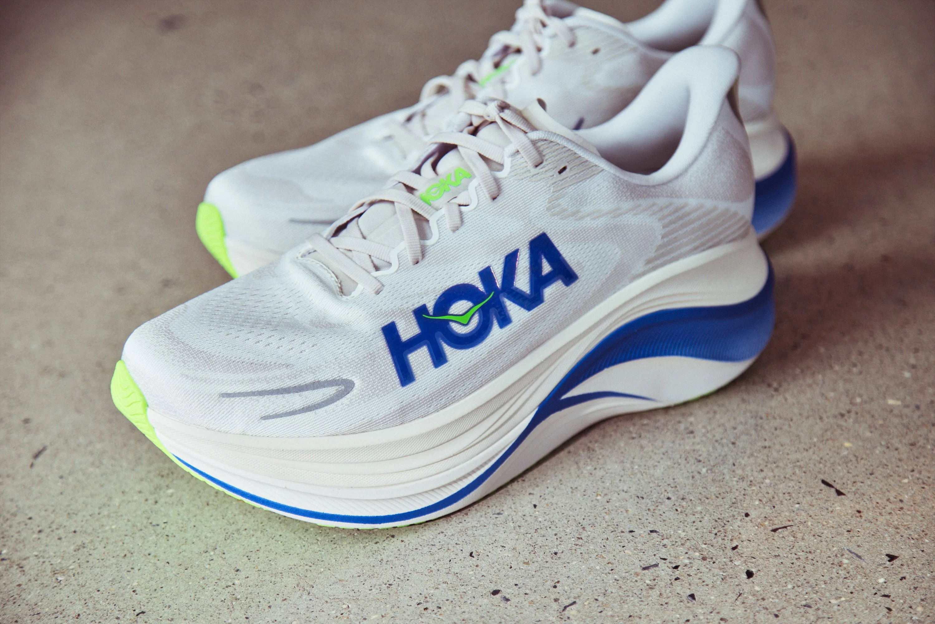 Hoka running shoes in white with blue accents and neon green soles, featuring a sleek design and prominent branding.