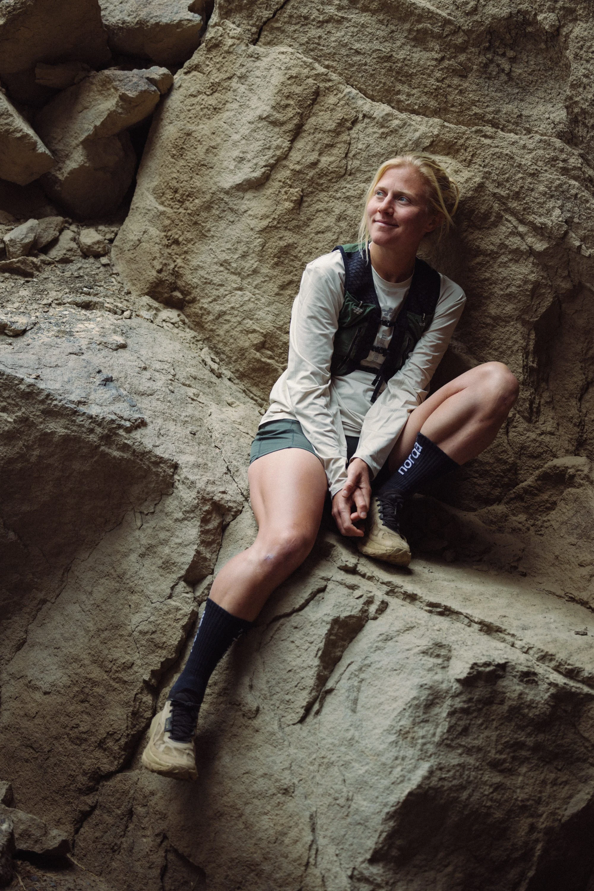A person sitting on a rocky surface, smiling and dressed in outdoor gear, surrounded by rough stone walls. A person sitting on a rocky surface, smiling and dressed in outdoor gear, surrounded by rough stone walls.