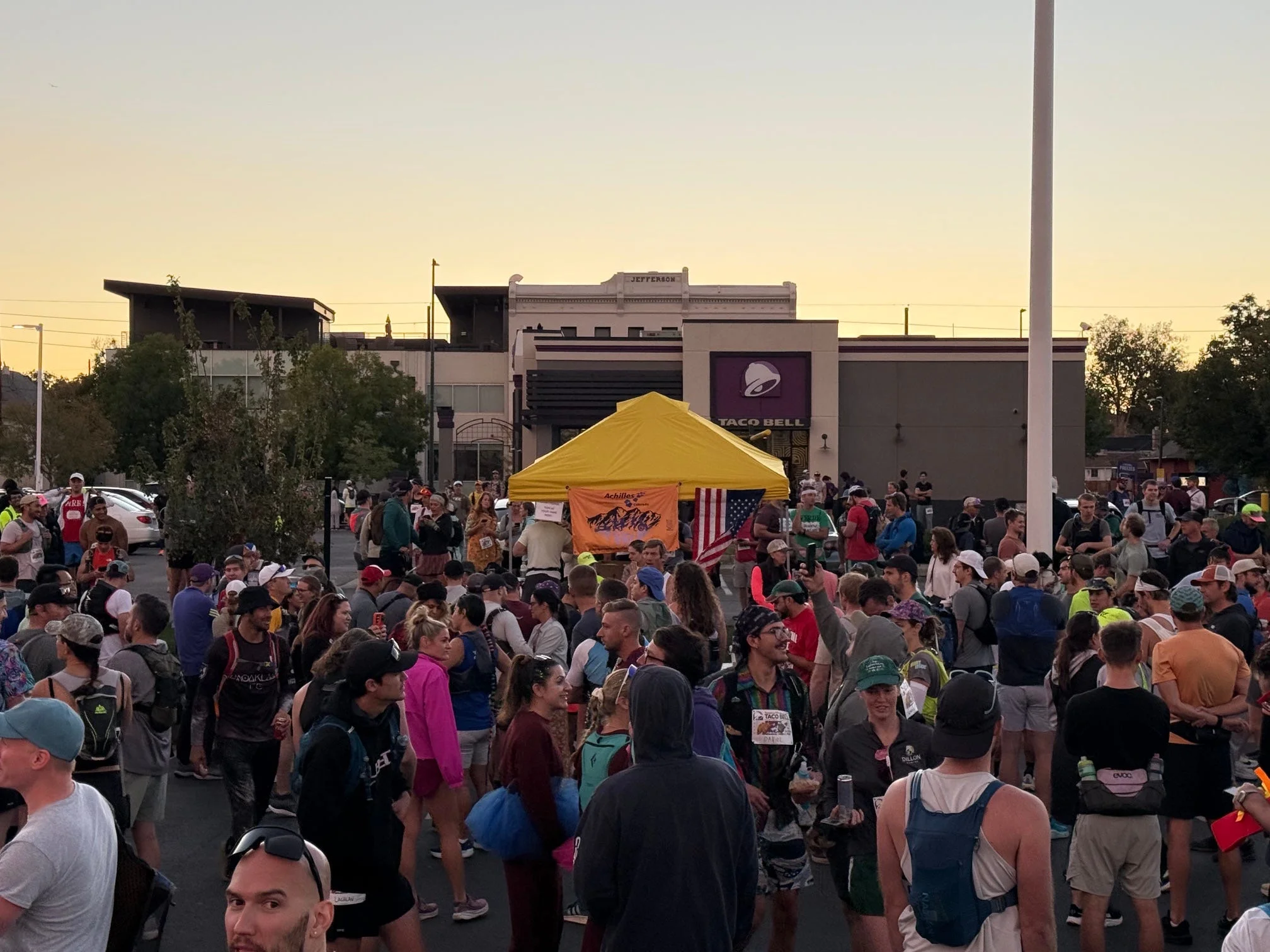A large crowd gathered around a yellow tent and a Taco Bell during an outdoor event at sunset. A large crowd gathered around a yellow tent and a Taco Bell during an outdoor event at sunset.