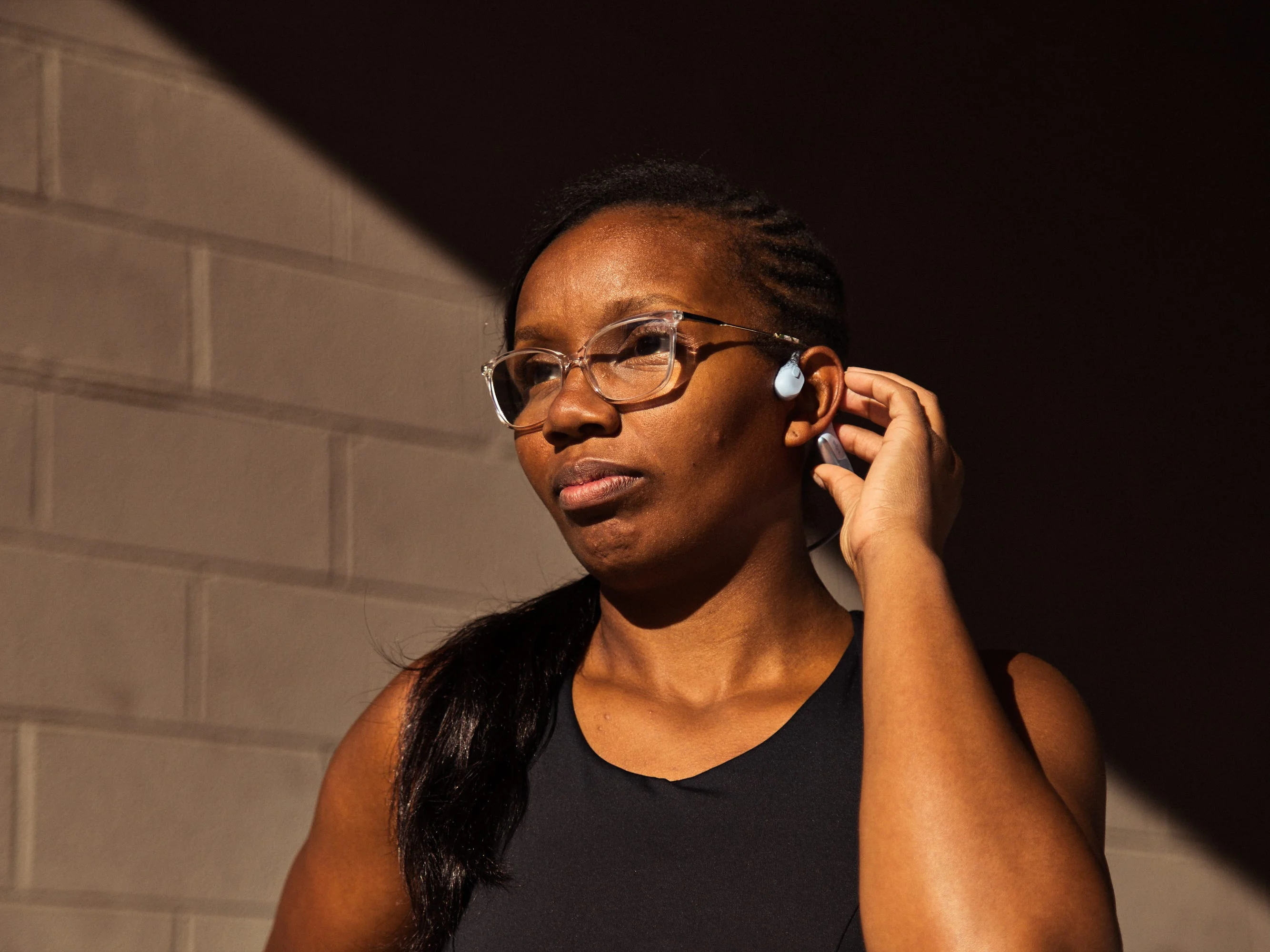 Woman with glasses adjusting her earbud while standing against a textured wall in warm light. Woman with glasses adjusting her earbud while standing against a textured wall in warm light.