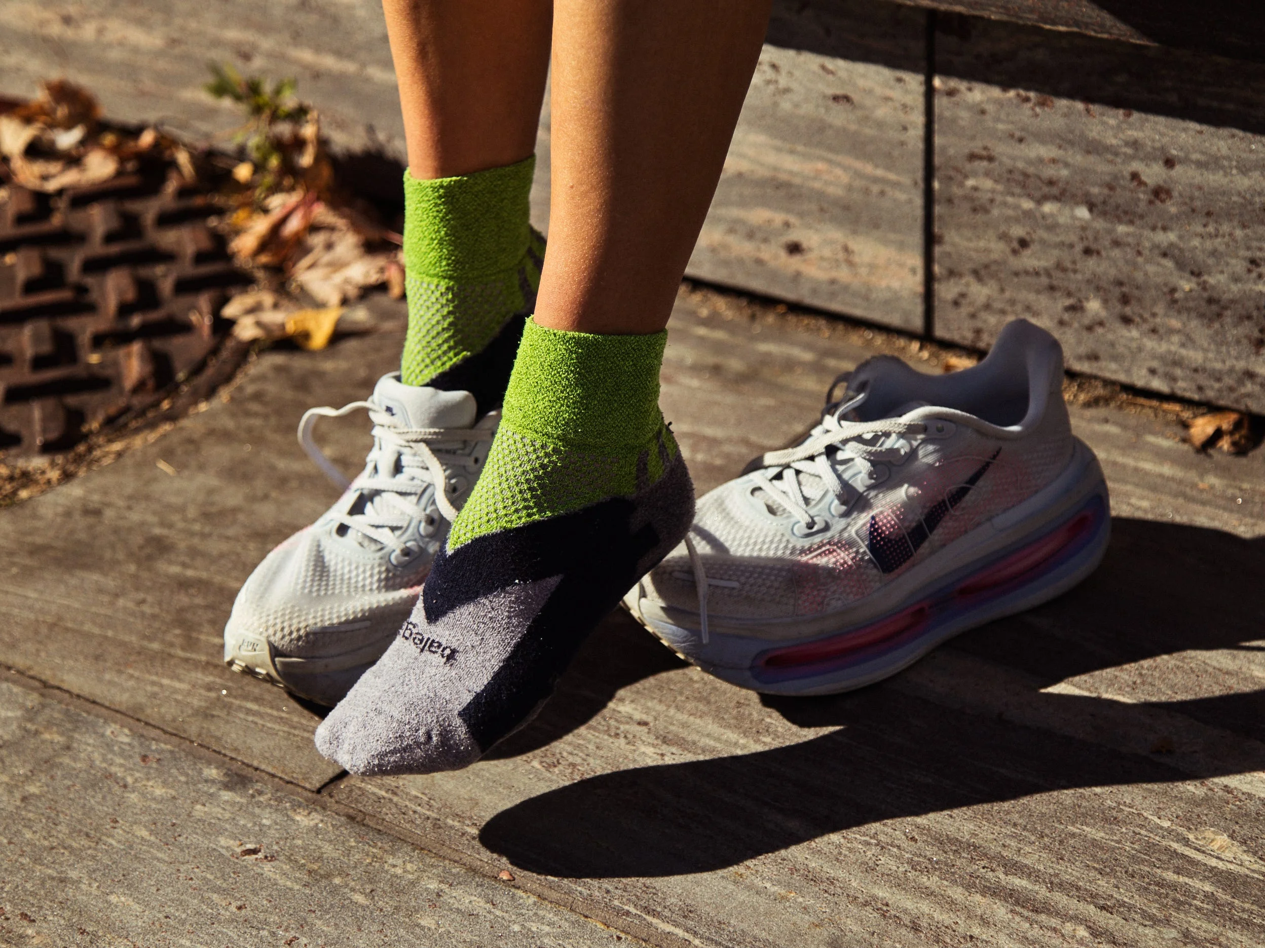 Feet wearing vibrant green and gray socks with sneakers resting on a textured surface, surrounded by autumn leaves.