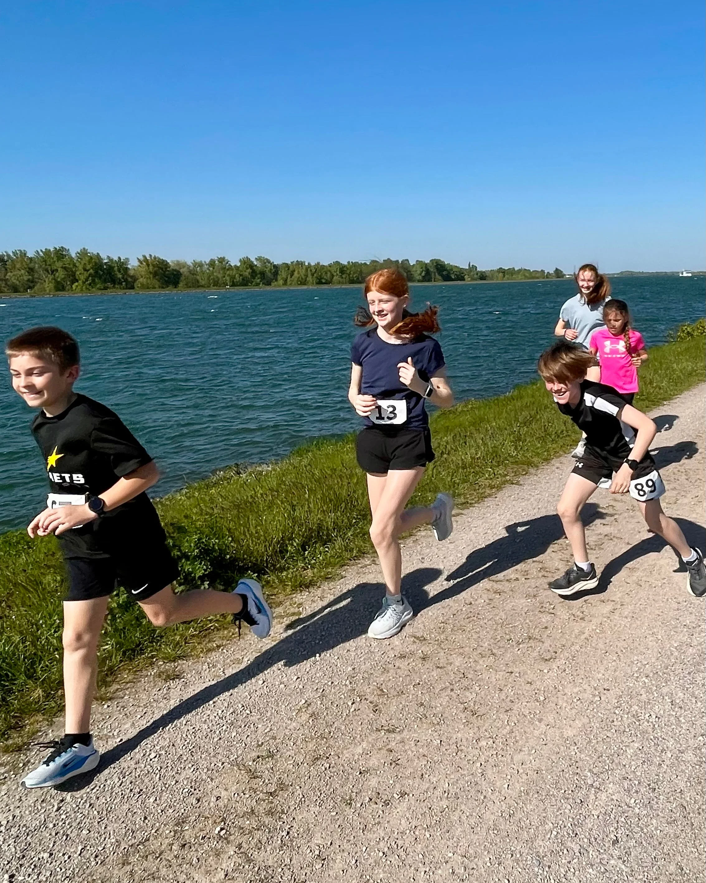 Children running along a pathway beside a body of water on a sunny day.