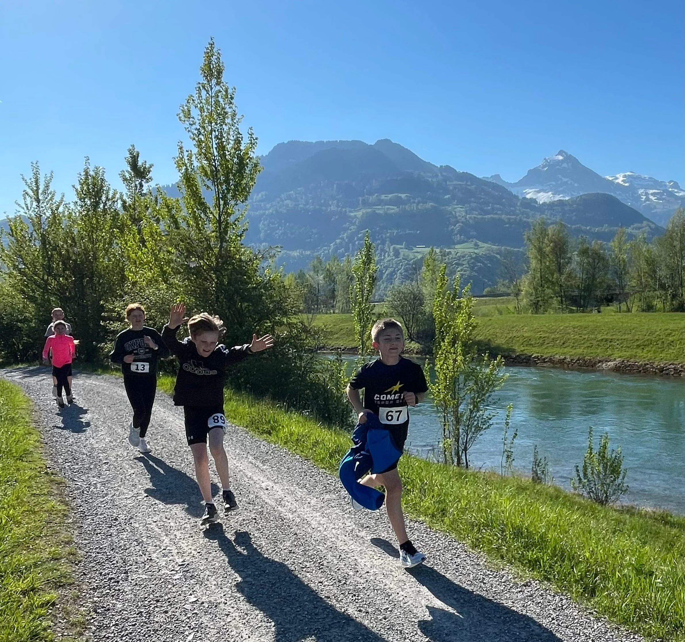 Children running on a gravel path by a river, surrounded by trees and mountains under a clear blue sky.
