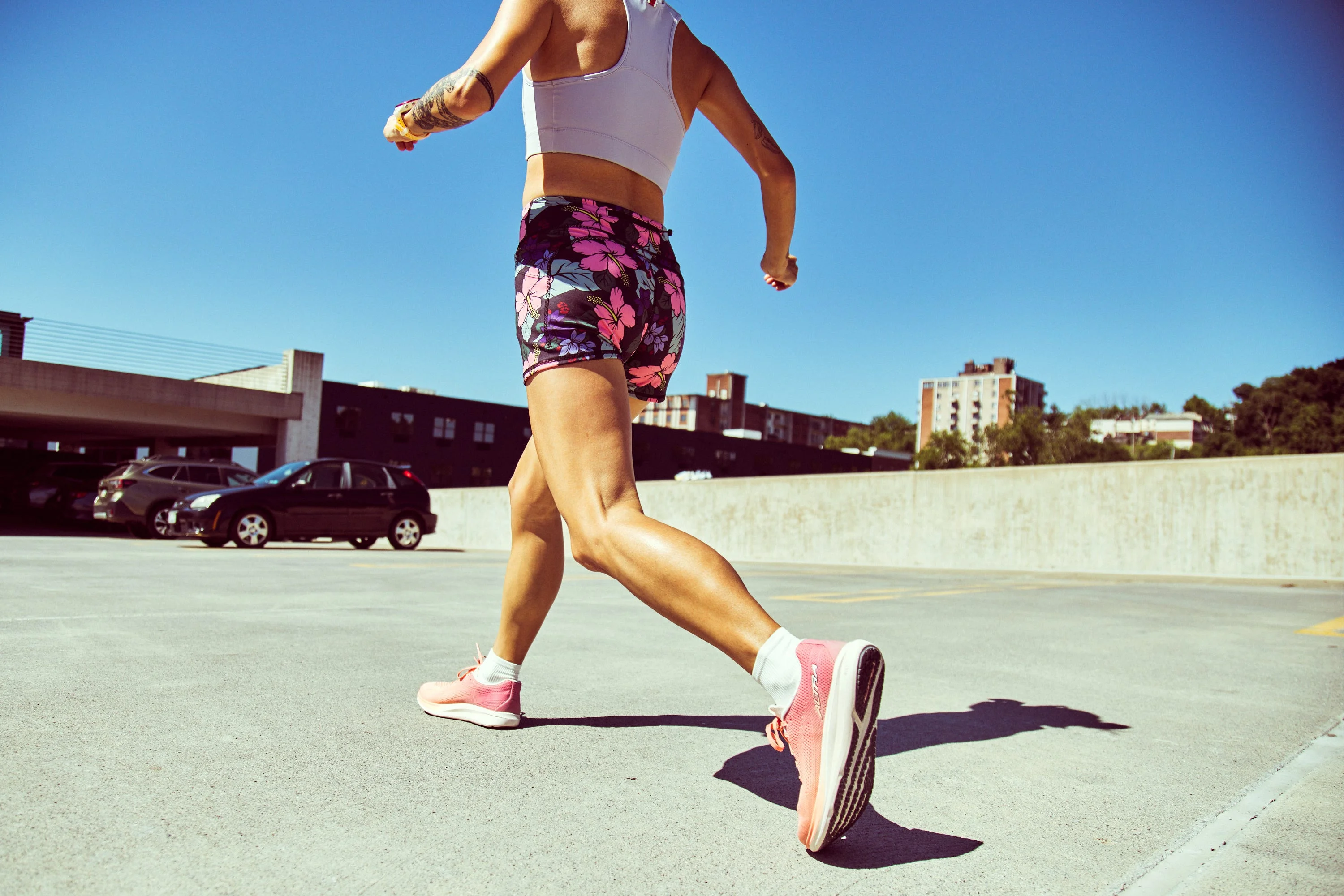 A person running in a parking area wearing a sports outfit and pink shoes under a clear blue sky.