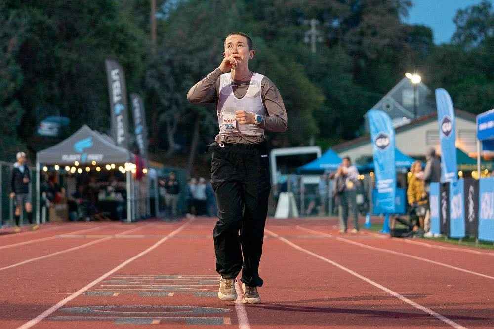 A runner on a track gestures with fingers to lips in a nighttime event, surrounded by spectators and tents.