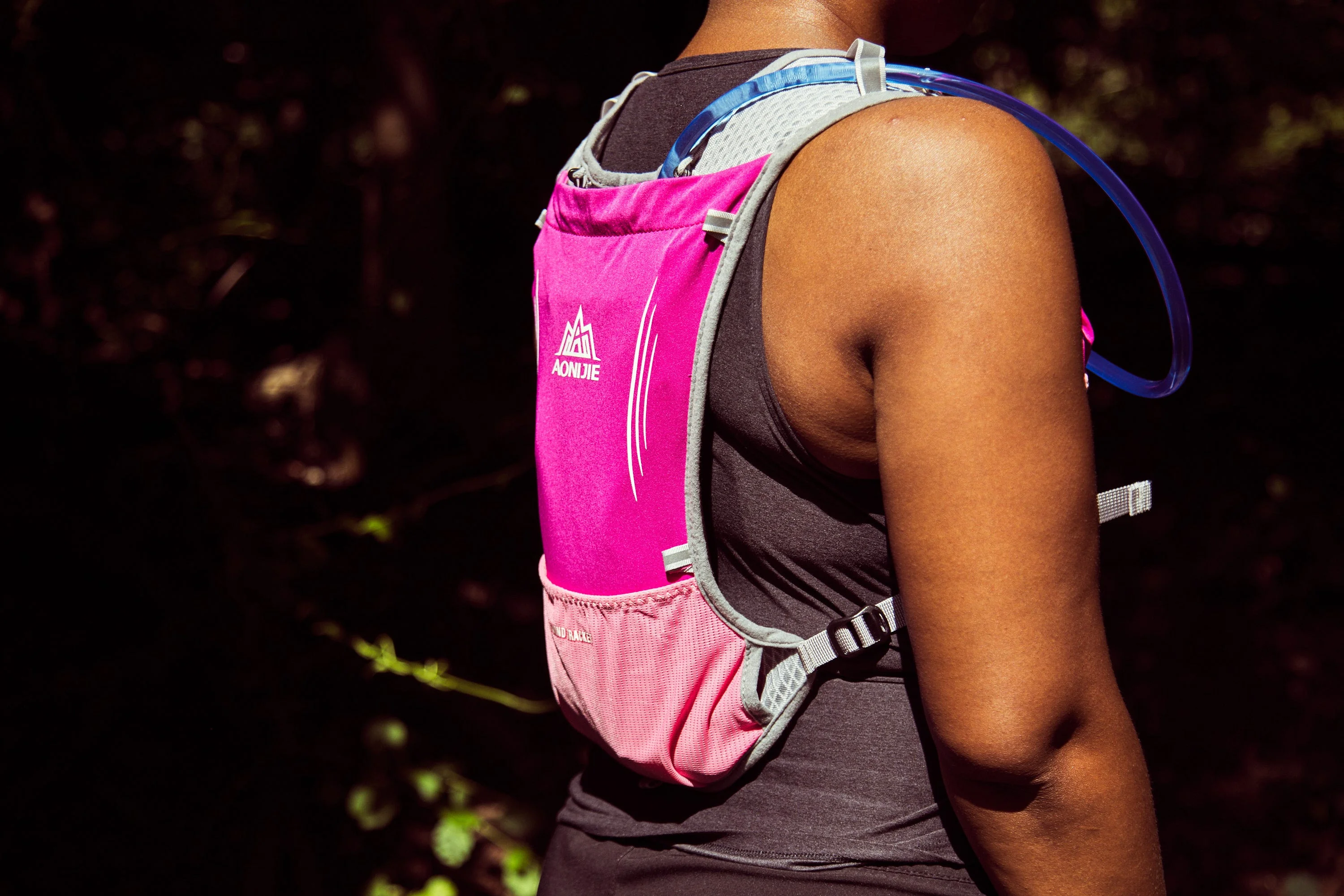 A close-up of a person wearing a pink hydration pack with a drinking tube in a forested setting.